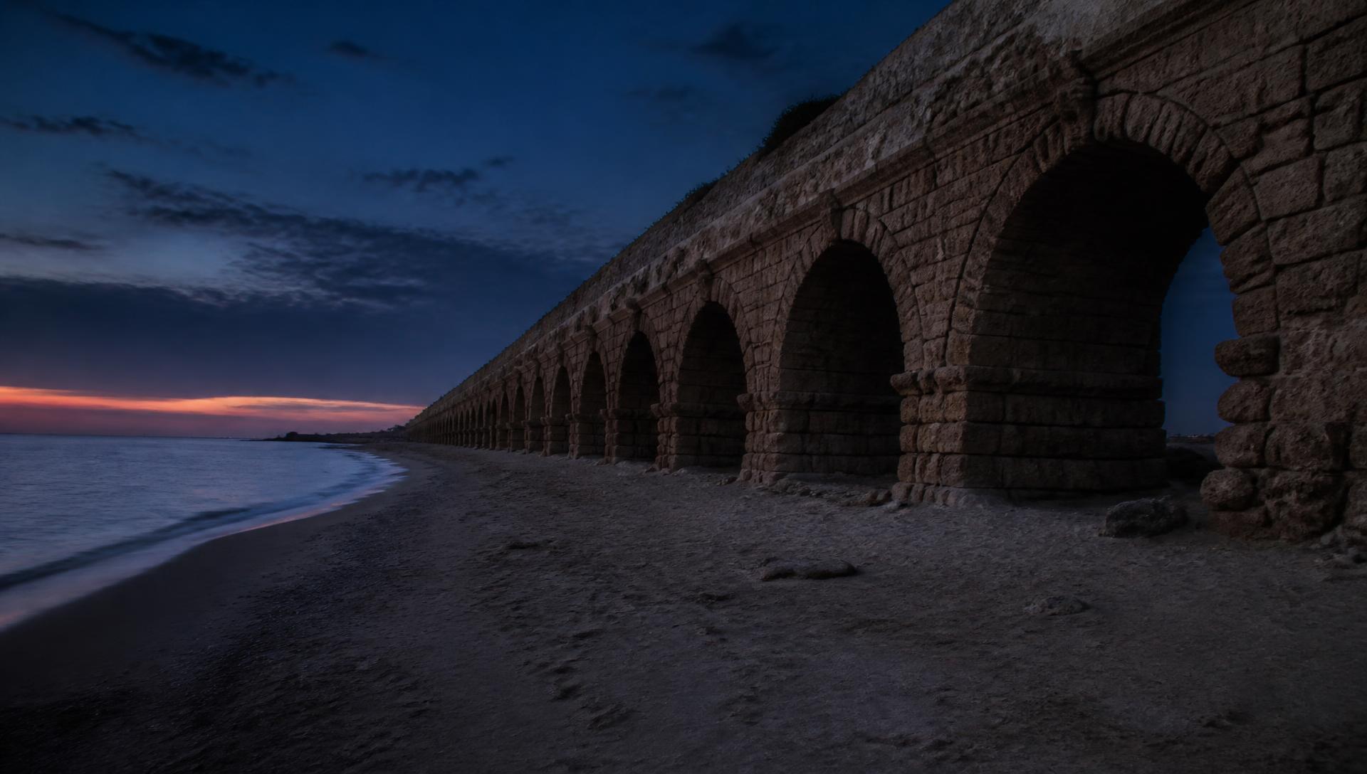 The arches of the ancient Roman aqueduct at Caesarea standing along the Mediterranean shore at dusk.